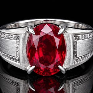 Puede incluir: Un anillo de tono plateado con una gran piedra preciosa de color rojo intenso y corte ovalado, sujeta por cuatro garras. La banda tiene un acabado de metal cepillado y está decorada con un patrón geométrico griego en ambos lados.