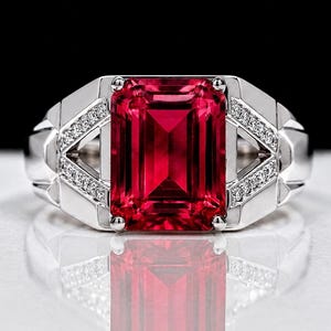 Op de afbeelding: Een zilverkleurige ring met een grote, rechthoekige, smaragdgeslepen, felrode edelsteen in het midden. De band is breed en metaalachtig met geometrische zijpanelen ingelegd met kleine, heldere, rondgeslepen witte steentjes aan beide kanten.