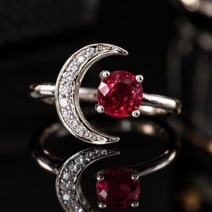 Peut inclure: Une bague argentée ornée d'un motif de croissant de lune incrusté de petites pierres claires et d'une unique pierre précieuse rouge foncé, ronde et facettée, fixée par quatre griffes à côté de la forme de lune sur une surface réfléchissante.