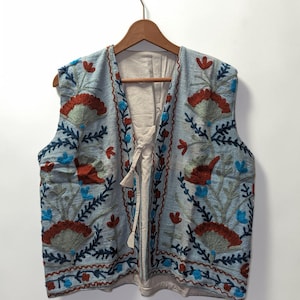 May include: A light blue sleeveless waistcoat featuring intricate embroidery in shades of rust orange, dark navy blue, and bright turquoise. The garment has a cream-coloured lining, a front tie closure, and a repeating floral pattern across the front.