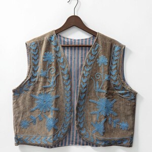 May include: A sleeveless waistcoat in a muted brown tone featuring intricate light blue floral embroidery along the front panels. The interior lining displays a vertical striped pattern in shades of blue, red, and white, visible at the open front.