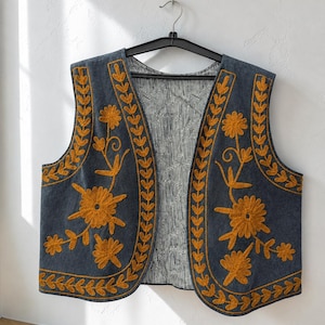 May include: A dark grey sleeveless vest featuring mustard yellow floral and leaf embroidery along the front edges. The interior lining displays a black and white abstract line pattern. The garment is displayed on a black plastic hanger.