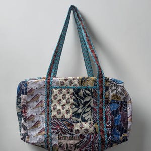 May include: A quilted tote bag featuring a patchwork design of various floral, bird, and paisley patterns in shades of blue, red, white, and black. The bag includes two long shoulder straps with a blue and white geometric print and blue piping.