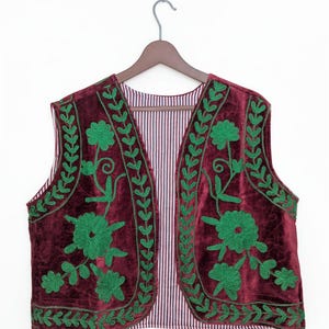 May include: A sleeveless waistcoat made of deep burgundy velvet fabric featuring vibrant green embroidered floral and vine patterns along the front panels. The interior lining is visible and displays a fine white and burgundy vertical striped pattern.