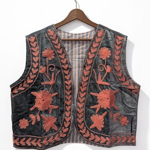 May include: A dark grey sleeveless vest featuring intricate coral-coloured floral and leaf embroidery along the front panels and hem. The garment is open-fronted and lined with a vertically striped fabric in shades of blue, white, and orange.