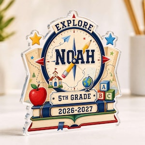 Peut inclure: Une plaque en acrylique transparent avec un motif de boussole, le nom NOAH, un crayon jaune, une pomme rouge, une école, un globe et des cubes alphabet. Le texte indique EXPLORE, NOAH, 5TH GRADE et 2026-2027, sur un fond de livres empilés.