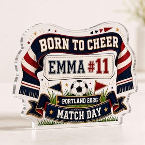May include: A clear acrylic plaque featuring a football theme with a navy and red scarf graphic. Text reads BORN TO CHEER, EMMA #11, PORTLAND 2026, and MATCH DAY. The design includes a football, green grass, and small flags on a white background.