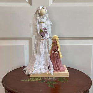 May include: Two handmade yarn dolls, one white with wings and a bouquet of purple flowers, the other pink with yellow hair, standing on a wooden base.