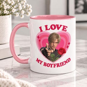 Puede incluir: Una taza de cerámica blanca con interior y asa de color rosa. El frente presenta un gráfico de corazón rosa que contiene la imagen de un hombre de cabello rubio sosteniendo una rosa roja, rodeado por el texto I LOVE MY BOYFRIEND en letras rosas.