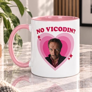 May include: A white ceramic mug with a pink interior and handle, featuring a pink heart graphic containing a portrait of a man with a serious expression. Above the heart, the text NO VICODIN? is printed in red, accompanied by small red star icons.