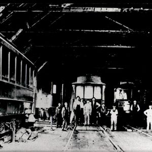 Peut inclure: Une photographie historique en noir et blanc montrant un groupe d'ouvriers debout à l'intérieur d'un grand dépôt ferroviaire. Plusieurs wagons d'époque sont positionnés sur des voies dans la structure sombre et caverneuse.