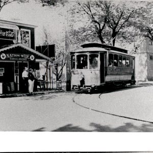 Peut inclure: Une photographie en noir et blanc montrant une scène de rue avec un tramway ancien sur des rails. À gauche, un bâtiment portant l'enseigne Nation-Wide Service Grocery affiche une publicité Coca-Cola. Des personnes se tiennent près de l'entrée.
