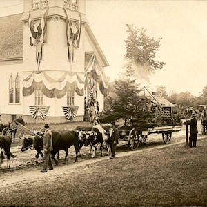 May include: A sepia-toned vintage photograph depicting a parade scene with a large white building decorated with draped bunting. A team of oxen pulls a wooden wagon carrying a small structure, while people in formal attire walk alongside the animals.