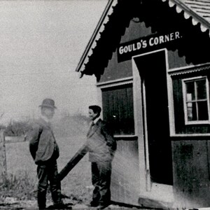 Peut inclure: Une photographie historique en noir et blanc montrant deux personnes debout devant une petite structure en bois avec une enseigne indiquant GOULD'S CORNER. Le bâtiment présente un toit à pignon avec des bordures décoratives et une fenêtre.