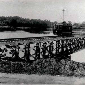 Peut inclure: Une photographie en noir et blanc d'un tramway électrique ancien traversant un long pont à chevalets en bois au-dessus d'une étendue d'eau calme. Le tramway est positionné sur des rails qui longent le pont, avec des poteaux électriques visibles.