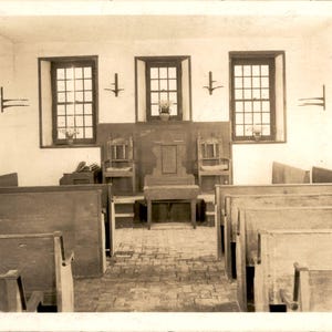 Peut inclure: Une photographie sépia d'un espace intérieur historique présentant des rangées de bancs en bois faisant face à une estrade centrale. L'estrade comprend une chaise en bois et une petite table, flanquées de deux chaises supplémentaires. Trois fenêtres à carreaux sont visibles.
