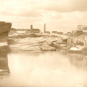 Peut inclure: Une photographie vintage sépia représentant un paysage côtier rocheux avec une mare résiduelle calme au premier plan. Au loin, une petite ville présente plusieurs bâtiments, une tour d'horloge et une haute cheminée sous un ciel nuageux.