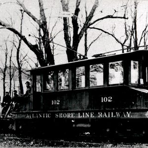 Peut inclure: Une photographie en noir et blanc d'un ancien tramway de l'Atlantic Shore Line Railway, numéroté 102, doté d'un chasse-pierres proéminent, de multiples fenêtres latérales et d'une perche de trolley, avec plusieurs personnes près de l'avant.