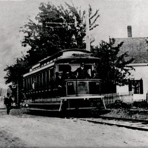 Peut inclure: Une photographie historique en noir et blanc d'un tramway électrique d'époque portant le numéro 19, circulant sur un chemin de terre à côté d'une clôture en bois et d'une maison. Le tramway présente de grandes fenêtres et un pantographe.