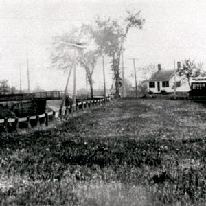 Peut inclure: Une photographie historique en noir et blanc montrant un tramway positionné près d'une petite maison blanche. Une clôture en bois longe un champ herbeux menant à un pont, avec des poteaux électriques et des arbres visibles en arrière-plan.