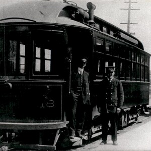 May include: A vintage black and white photograph of a dark-colored electric streetcar with the number 18 visible on the front. Two uniformed operators stand near the open entrance of the rail vehicle, which features large windows and a roof.