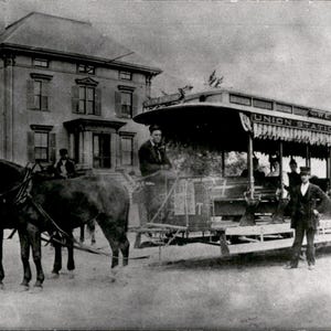 Peut inclure: Une photographie en noir et blanc d'un tramway tiré par deux chevaux au premier plan. Le tramway présente des côtés ouverts, des bancs et une inscription indiquant Union Station & West End. Plusieurs personnes sont assises et debout.