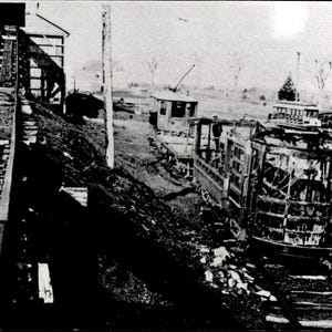 Peut inclure: Une photographie historique en noir et blanc montrant un tramway ouvert endommagé reposant sur des rails. Une structure en bois se dresse sur la gauche avec une personne à proximité. En arrière-plan, un autre tramway et des véhicules ferroviaires.