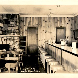 Peut inclure: Une carte postale vintage sépia montrant l'intérieur de Symonds à Wells Beach, Maine. La scène présente une cheminée en pierre, des murs lambrissés, un long comptoir de service avec plusieurs tabourets et plusieurs tables avec des chaises.