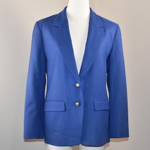 May include: A solid royal blue blazer featuring a notched lapel, long sleeves, and two gold-toned metal buttons at the center front closure. The garment includes two horizontal flap pockets positioned at the lower front on each side.
