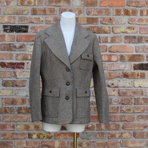 May include: A brown and white houndstooth blazer featuring a notched lapel, three dark buttons down the front, two large flap pockets at the waist, and a single smaller flap pocket on the upper left chest, displayed on a white mannequin torso.
