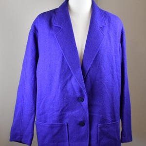 May include: A vibrant purple blazer featuring a notched lapel, long sleeves, and two dark buttons down the front. The garment includes two large patch pockets at the lower front and is displayed on a mannequin to show its structured shape.