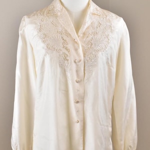 Puede incluir: Una blusa de manga larga color crema con botones y un intrincado patrón floral bordado en el cuello y la zona del pecho. La prenda tiene un cuello en punta, mangas largas con puños abotonados y un tejido liso con un acabado ligeramente brillante.