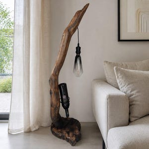 Pode incluir: Um candeeiro de pé feito de um ramo de madeira flutuante natural castanho-escuro, com um candeeiro suspenso preto, uma lâmpada em forma de gota e um suporte integrado para uma garrafa de vinho, assente numa base de madeira maciça.