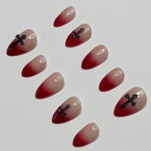 Peut inclure: Un ensemble de dix faux ongles en forme d'amande avec un dégradé passant d'une base nude à une pointe rouge foncé. Trois des ongles sont ornés d'un petit emblème en forme de croix noire texturée, placé au centre de la surface de l'ongle.