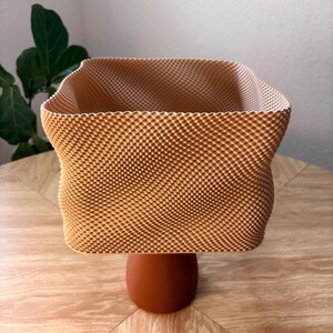May include: A square-shaped vessel with a textured, light tan, grid-like exterior pattern. The container features a wide, open top with slightly curved sides and is supported by a solid, dark orange, circular pedestal base with a fine, dotted texture.