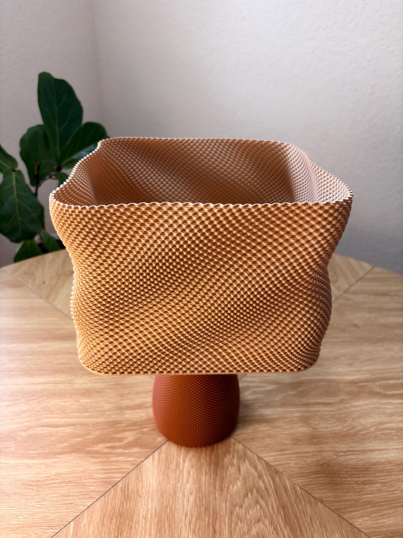 May include: A square-shaped vessel with a textured, light tan, grid-like exterior pattern. The container features a wide, open top with slightly curved sides and is supported by a solid, dark orange, circular pedestal base with a fine, dotted texture.