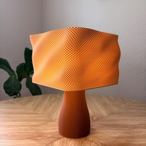 May include: A table lamp with a vibrant orange, wavy, square-shaped shade featuring a fine, textured honeycomb pattern. The base is a solid, darker orange, cylindrical shape that also displays a consistent, fine-textured grid pattern throughout.