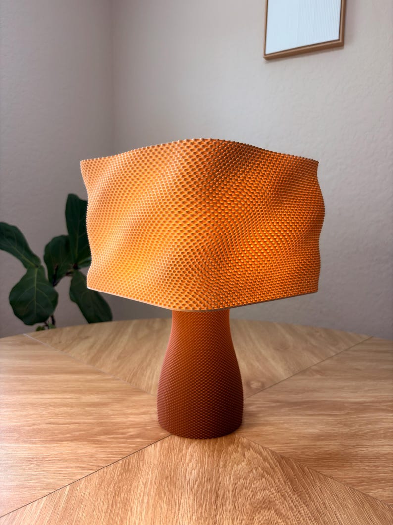 May include: A table lamp with a vibrant orange, wavy, square-shaped shade featuring a fine, textured honeycomb pattern. The base is a solid, darker orange, cylindrical shape that also displays a consistent, fine-textured grid pattern throughout.