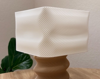 Pode incluir: Um candeeiro de mesa com um abajur quadrado, branco e texturizado com um padrão de grelha fina. A base é um suporte cilíndrico de cor castanha clara com uma textura de superfície ondulada e canelada que apresenta um padrão de grelha fino.