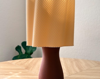 Pode incluir: Um candeeiro de mesa com uma base texturizada em forma de ampulheta castanho-escura e um abajur cilíndrico com bordas onduladas em amarelo claro. Toda a superfície da base e do abajur apresenta um padrão de pontos fino e consistente.