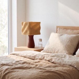 May include: A table lamp with a textured, wavy mustard yellow shade and a dark brown, tapered base. The lamp sits on a light wood nightstand next to a clear glass carafe filled with water, positioned beside a bed with beige linen bedding.