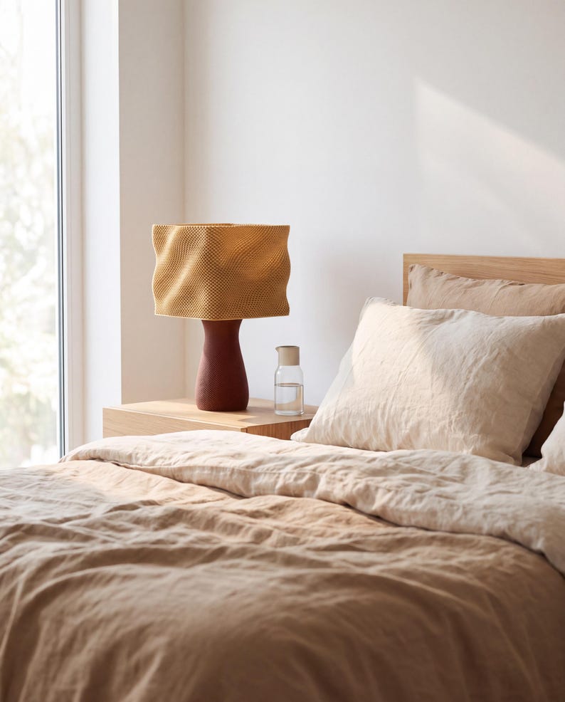 May include: A table lamp with a textured, wavy mustard yellow shade and a dark brown, tapered base. The lamp sits on a light wood nightstand next to a clear glass carafe filled with water, positioned beside a bed with beige linen bedding.