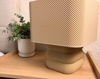Pode incluir: Um candeeiro de mesa bege com um abajur quadrado texturizado e uma base quadrada arredondada e empilhada. O candeeiro apresenta um padrão de pontos consistente em toda a sua superfície, criando uma aparência uniforme.