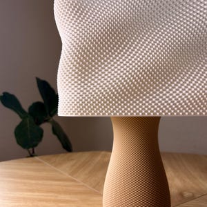 May include: A table lamp featuring a white rectangular shade with a textured, grid-like surface pattern. The base is a tan, hourglass-shaped stand that also displays a fine, uniform dotted texture across its entire exterior surface.