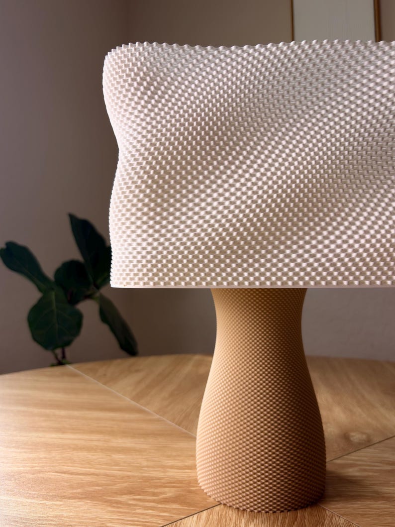 May include: A table lamp featuring a white rectangular shade with a textured, grid-like surface pattern. The base is a tan, hourglass-shaped stand that also displays a fine, uniform dotted texture across its entire exterior surface.
