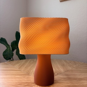 May include: A table lamp with a textured, orange, rectangular shade featuring a wavy, undulating surface pattern. The base is a solid, tapered, dark orange cylinder with a fine, uniform, grid-like texture that matches the shade's intricate design.