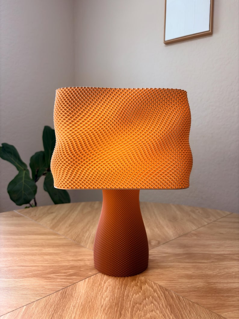 May include: A table lamp with a textured, orange, rectangular shade featuring a wavy, undulating surface pattern. The base is a solid, tapered, dark orange cylinder with a fine, uniform, grid-like texture that matches the shade's intricate design.
