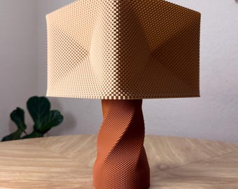 May include: A table lamp featuring a square, light beige shade with a textured, grid-like surface and a central indentation on each side. The base is a cylindrical, dark orange stand with a spiraling, twisted design and a matching grid texture.