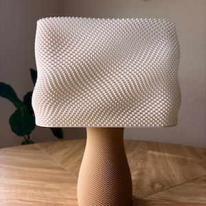 May include: A table lamp featuring a rectangular, off-white lampshade with a textured, grid-like surface pattern. The base is a tan, cylindrical pedestal with a tapered middle section, also displaying a fine, uniform, textured grid pattern.