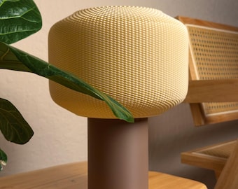Pode incluir: Um candeeiro de mesa com uma base cilíndrica castanha e um abajur arredondado, texturizado, amarelo-pálido com um padrão de grelha fina. A palavra "solenne." está impressa numa fonte cursiva pequena e escura acima do abajur.